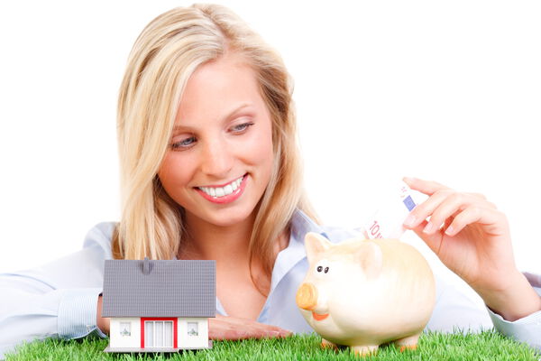 Ihr Immobilien-Tipp wird belohnt – Kurzurlaub, Gutscheine oder Bargeld Junge Frau steckt Geldschein in ein Sparschwein, neben einem Miniaturhaus auf einer grünen Wiese – Symbol für Sparen und Eigenheimfinanzierung.